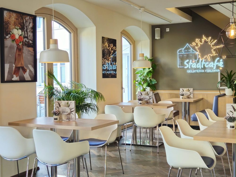 Gastraum des Stadtcafés mit hellen Tischen, Pflanzen und dem Leuchtschriftzug Stadtcafé Gelateria Italiana